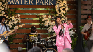 Woman in pink suit singing on stage with Water Park sign on the wooden back of stage.